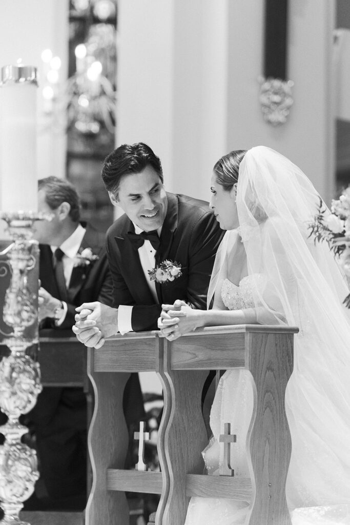 Black-and-white moment of the bride and groom leaning toward one another during the ceremony, sharing a quiet smile at the pews.