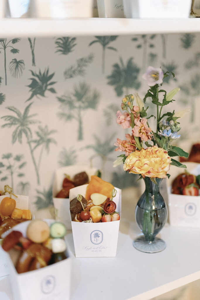 Late-night wedding snacks displayed in custom paper cones beside a small floral arrangement on a patterned bar shelf.