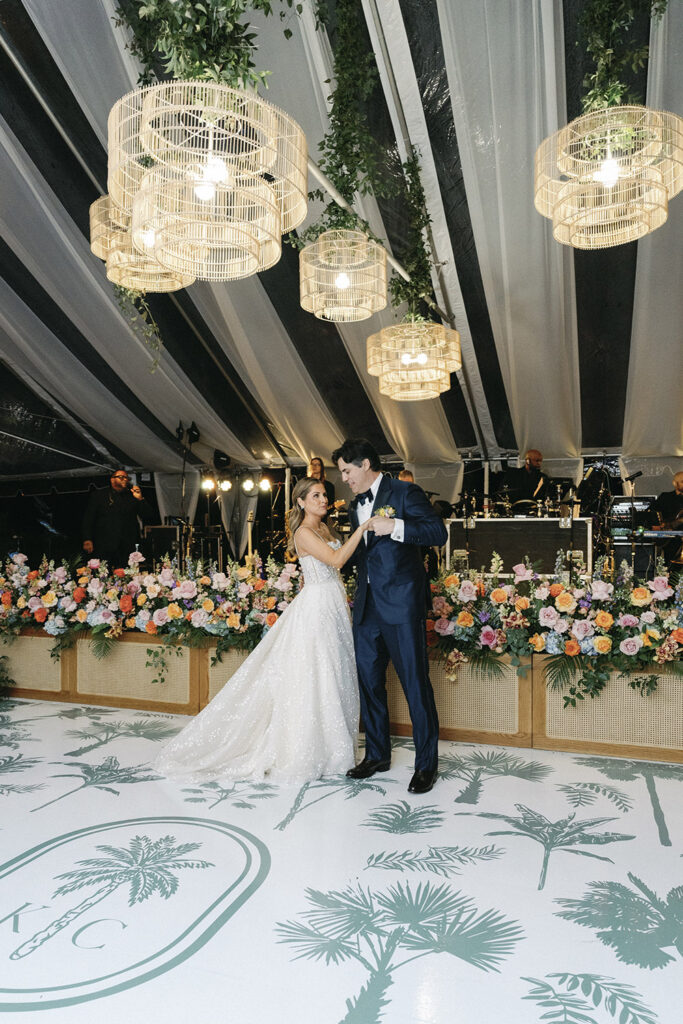 Bride and groom sharing their first dance beneath woven pendant lights and a lush floral stage backdrop inside the reception tent.