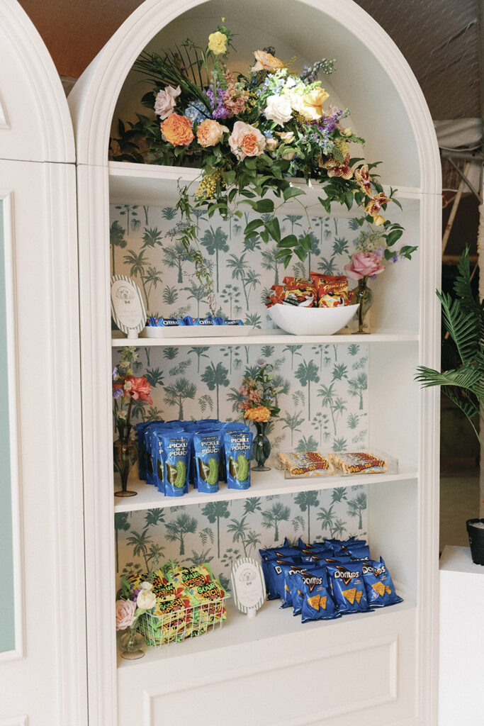 Curated snack wall featuring chips, candy, and late-night treats styled with palm-print wallpaper and floral accents.