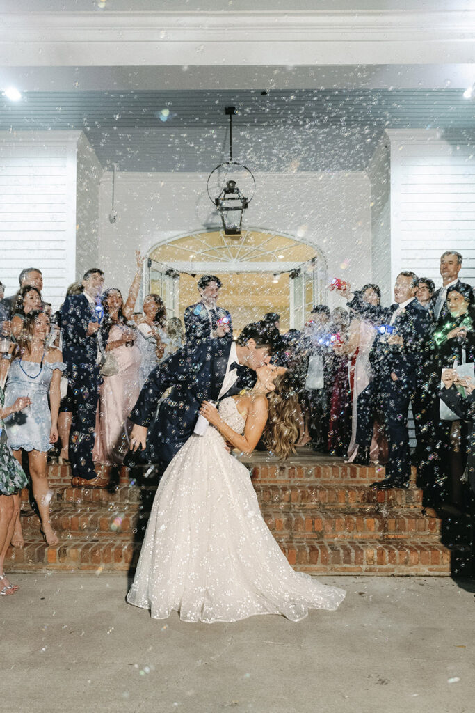 Bride and groom sharing a dramatic dip kiss during their sparkler-style bubble exit, surrounded by cheering guests on the venue steps at night.