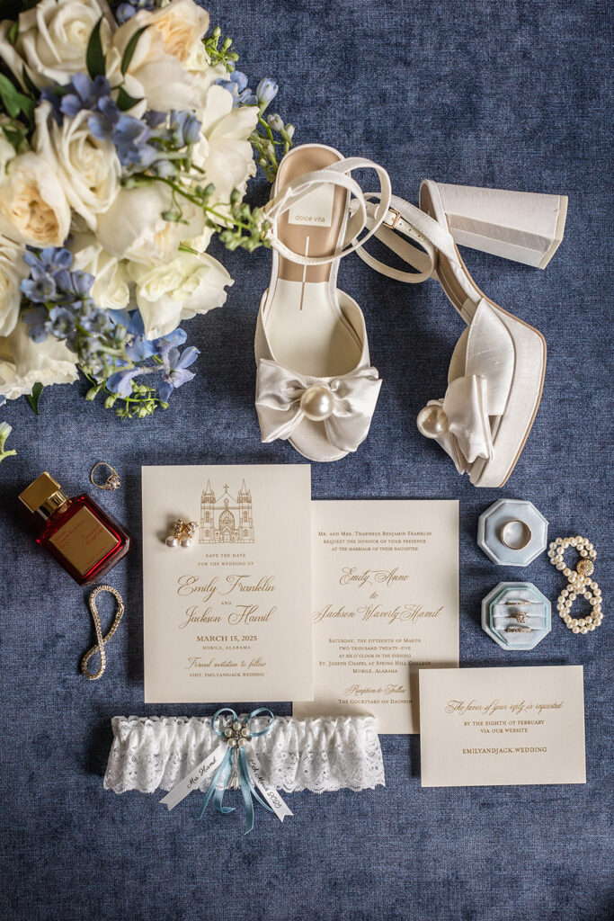 Flat lay of wedding details including invitation suite, shoes, jewelry, perfume, and garter on blue fabric.