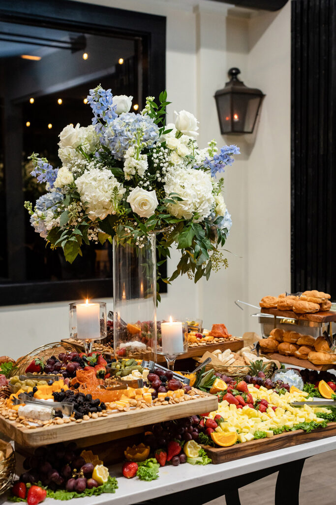 Reception food display with charcuterie boards, fruit, cheeses, and floral centerpiece with candles.