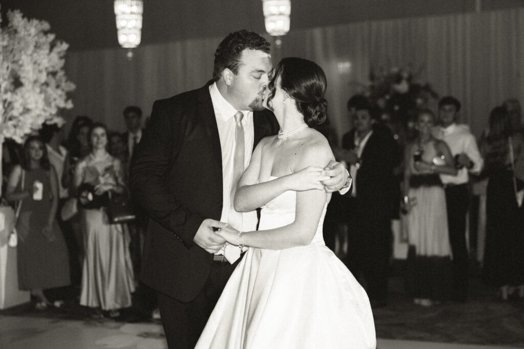 Newlywed couple sharing their first dance and a kiss on the reception dance floor surrounded by guests.