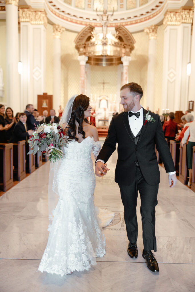 Newlyweds walk hand in hand down the church aisle as guests stand and applaud.