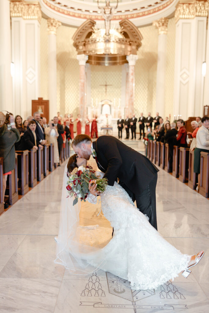 The groom dips the bride for their first kiss in the church aisle as guests stand and watch from the pews.