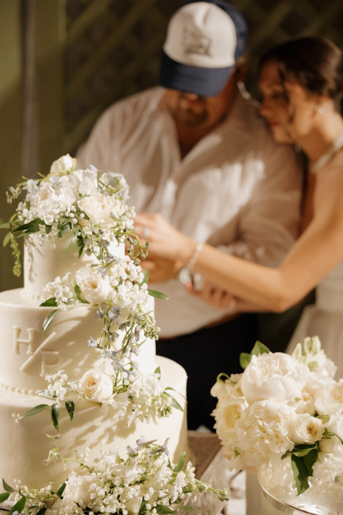 .Three-tier white wedding cake decorated with fresh white flowers while the couple cuts the cake in the background.