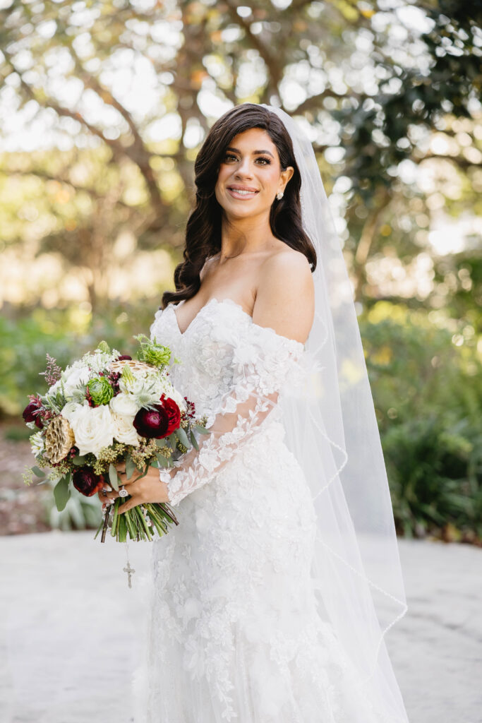 Outdoor bridal portrait of the bride in a lace off-the-shoulder gown and veil, holding a bouquet of white, red, and green florals against a soft garden backdrop.