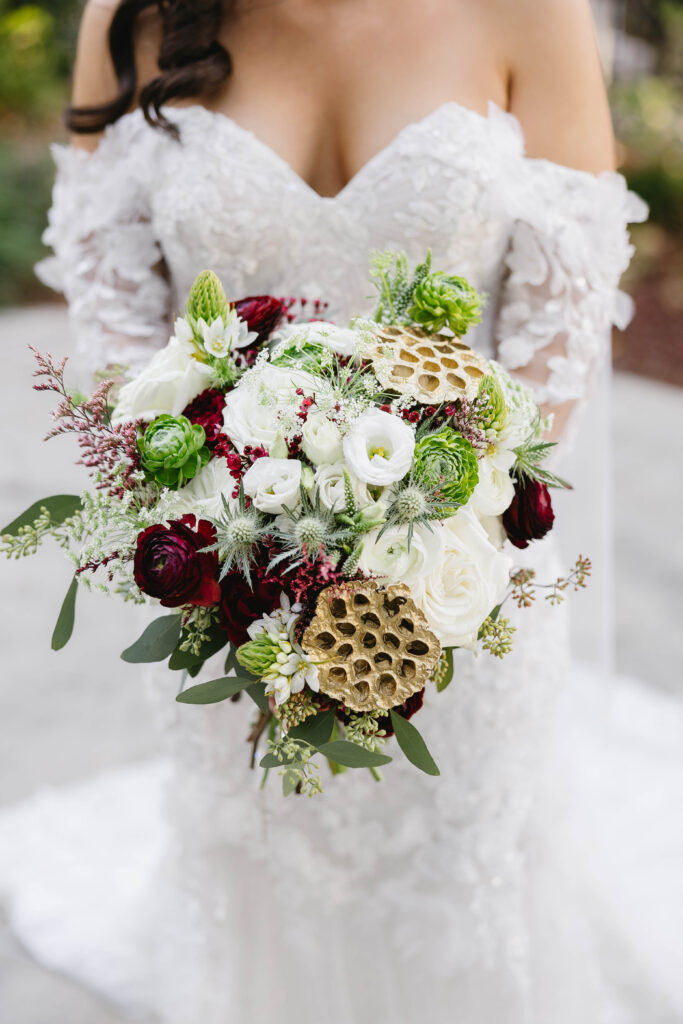 Detailed bouquet of white and deep red blooms with greenery and gold lotus pods against the bride’s lace gown.