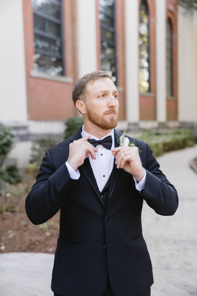 Groom adjusts his black bow tie during an outdoor portrait in front of the church.