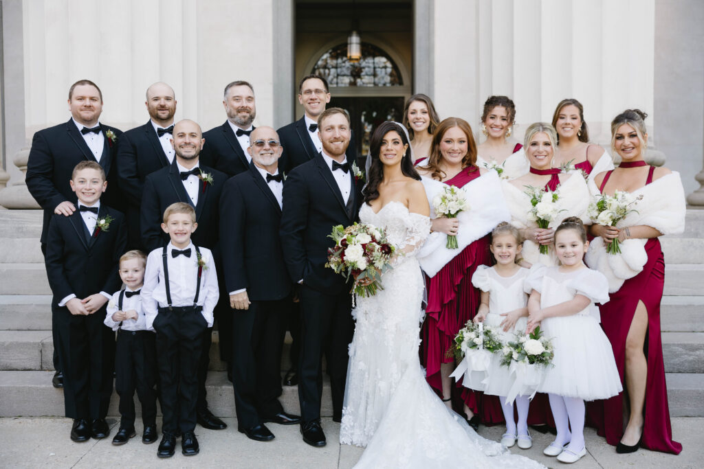Formal wedding party portrait on church steps with the bride and groom centered, surrounded by bridesmaids in red dresses, groomsmen in black tuxedos, flower girls in white, and young ring bearers.