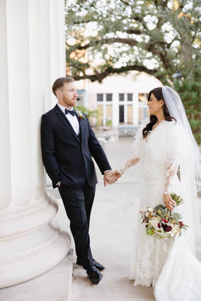 Bride and groom hold hands between grand white columns, smiling at one another during an outdoor portrait.