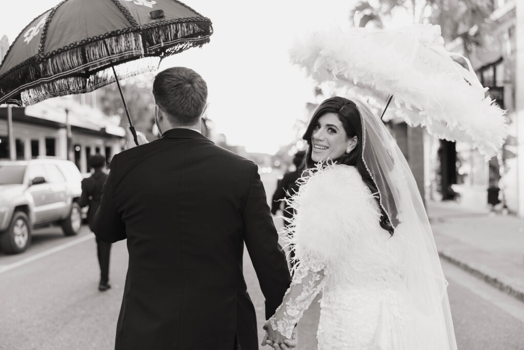 Black-and-white photo of the bride and groom walking down a city street holding decorative parasols, the bride smiling back over her shoulder.