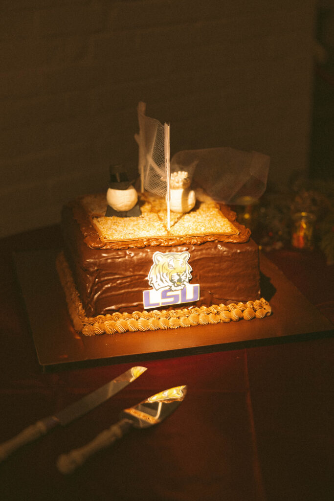 Groom’s cake decorated with an LSU logo and small bride-and-groom figurines on top, displayed in warm candlelight.