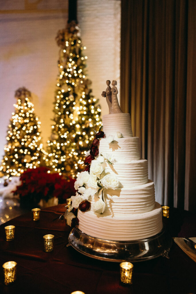 Four-tier white wedding cake adorned with cascading red and white flowers, set before glowing Christmas trees.