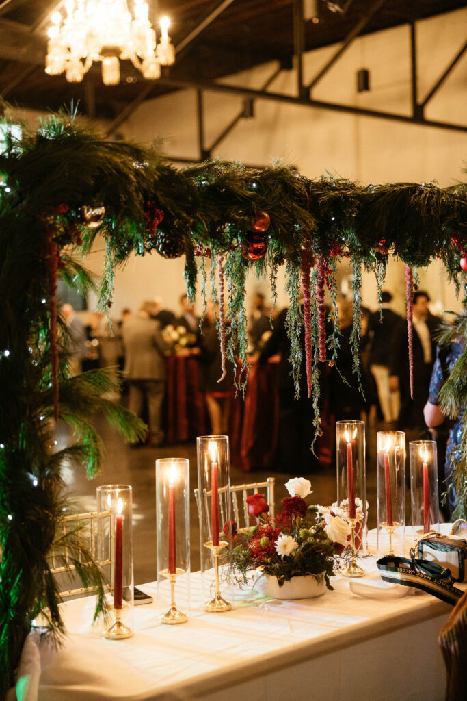 Reception table styled with tall red taper candles in glass holders, lush greenery overhead, and guests mingling in the softly lit background.