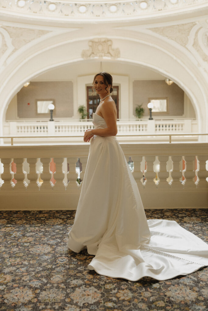 Bride in a strapless satin wedding gown posing beneath an ornate historic balcony inside the venue.