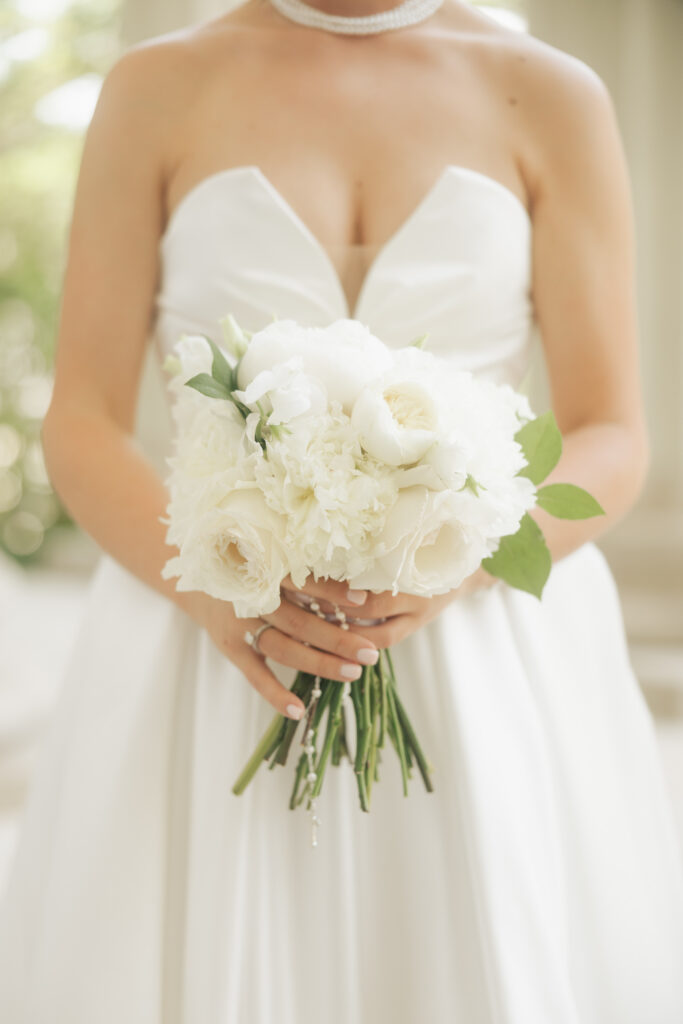 Close-up of bride holding a bouquet of white roses and peonies while wearing a pearl necklace and strapless gown.
