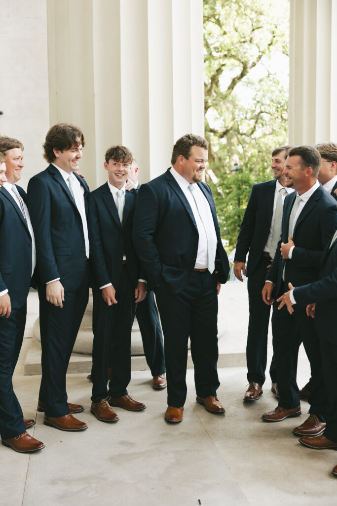 Group of groomsmen in navy suits laughing together outside a columned wedding venue before the ceremony.