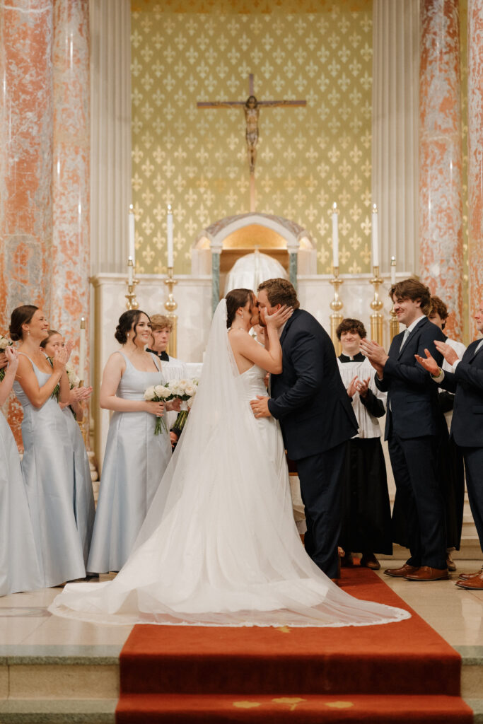 Bride and groom sharing their first kiss at the altar as wedding party members applaud.
