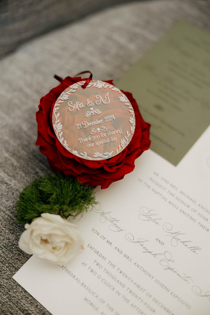Close-up of a personalized clear ornament reading “Sofia & AJ, 21 December 2024” resting on a red rose atop the wedding invitation.
