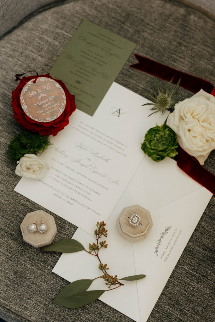 Flat lay of wedding invitation suite with pearl earrings, diamond engagement ring, white and red florals, greenery, and a personalized “Sofia & AJ” ornament dated December 21, 2024.