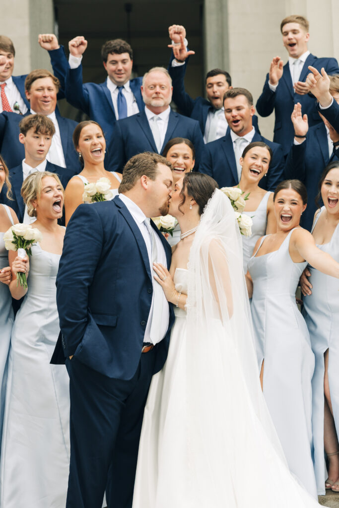 Wedding party cheering as the bride and groom kiss during a joyful group portrait outside the church.