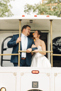 Newlywed couple smiling at each other while riding on the back platform of a vintage white trolley.