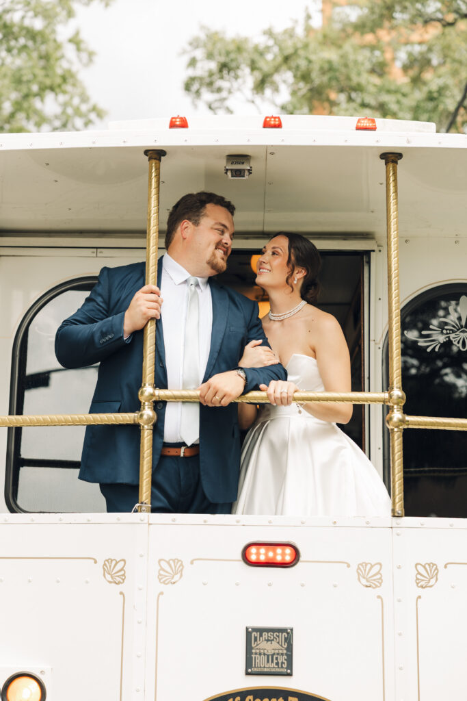 Newlywed couple smiling at each other while riding on the back platform of a vintage white trolley.
