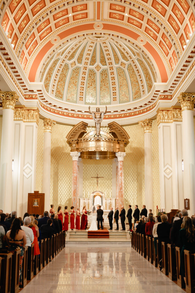Wide view of the ornate church interior during the ceremony, with the bride and groom at the altar beneath a grand domed ceiling and guests seated in the pews.