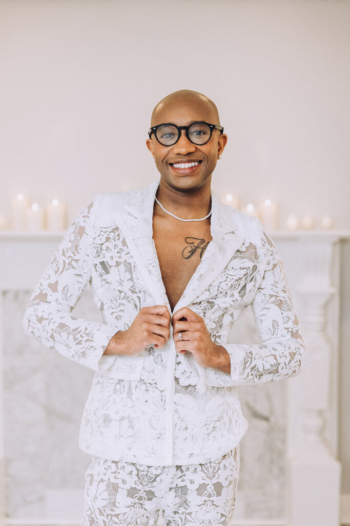 Person wearing a white lace wedding suit jacket and pearl necklace smiling while getting ready before the ceremony.