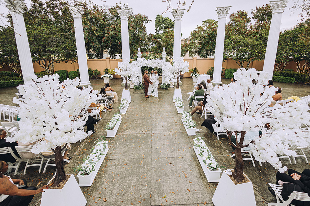 Outdoor wedding ceremony set beneath tall white columns with a lush white floral arch and aisle lined with decorative white trees.