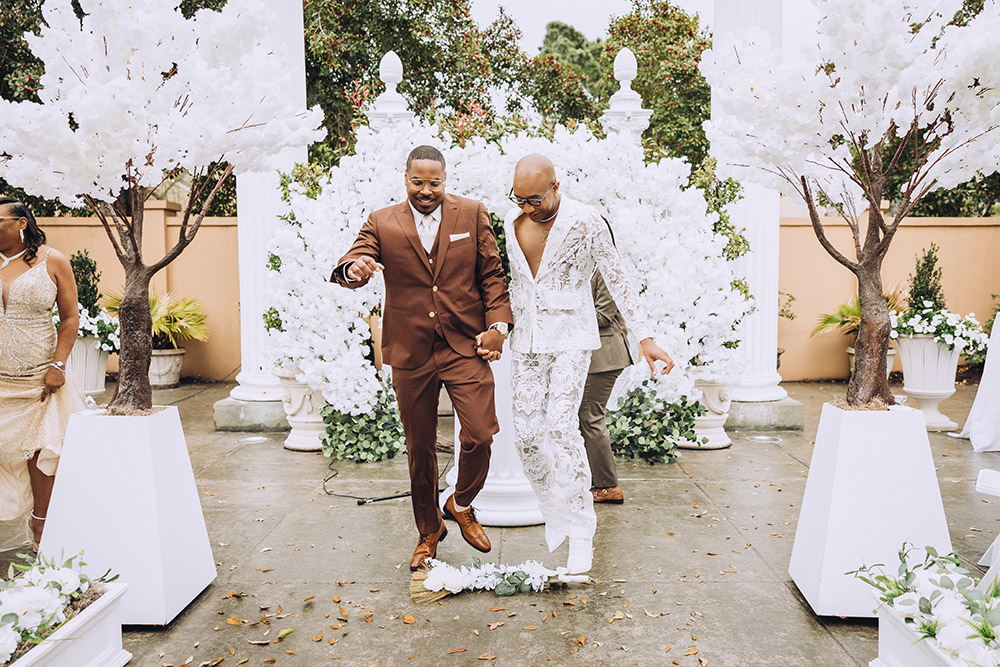 Newlywed couple walking down the ceremony aisle beneath white floral trees and columns after exchanging vows.