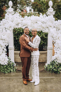 Newlywed couple embracing beneath a white floral ceremony arch during their outdoor wedding celebration.