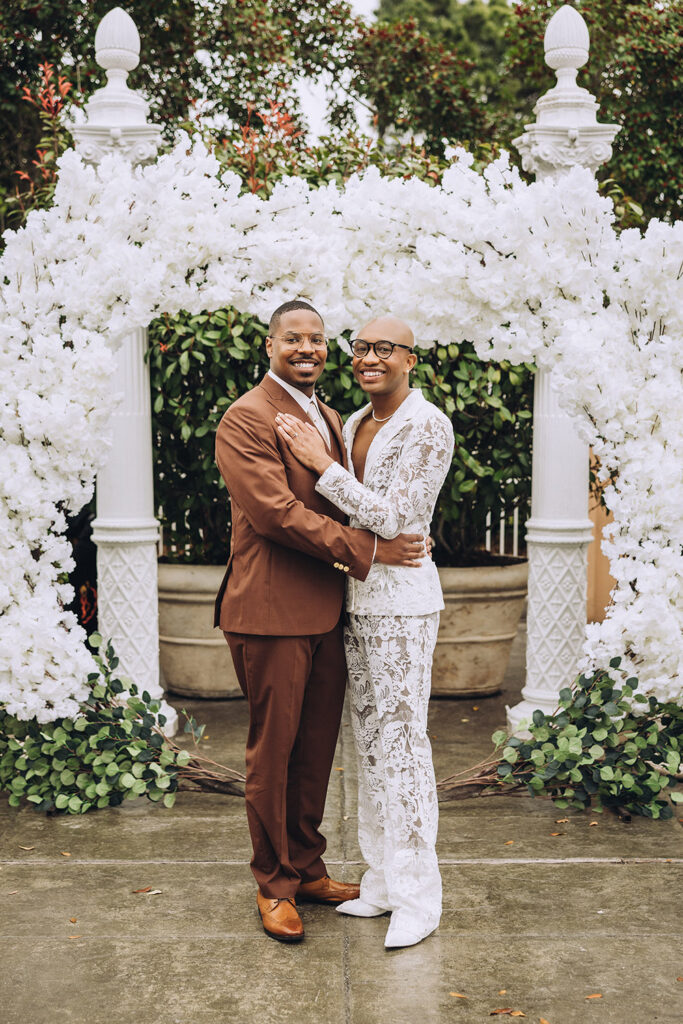Newlywed couple embracing beneath a white floral ceremony arch during their outdoor wedding celebration.