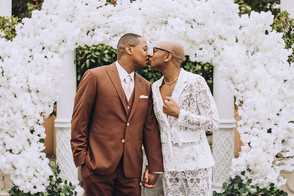 Newlywed couple sharing a kiss beneath a lush white floral ceremony arch.