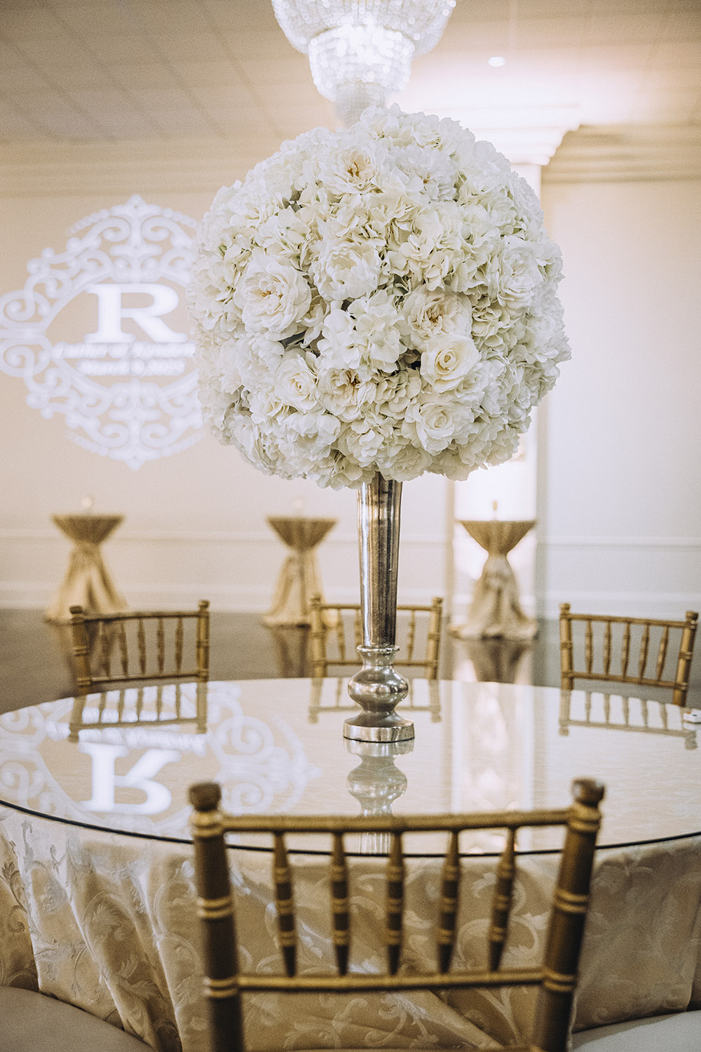 Elegant reception table with gold chairs and a tall white floral centerpiece beneath a chandelier.