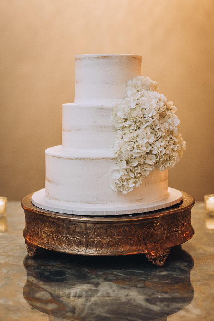 Three-tier white wedding cake decorated with cascading white hydrangeas displayed on an ornate stand.