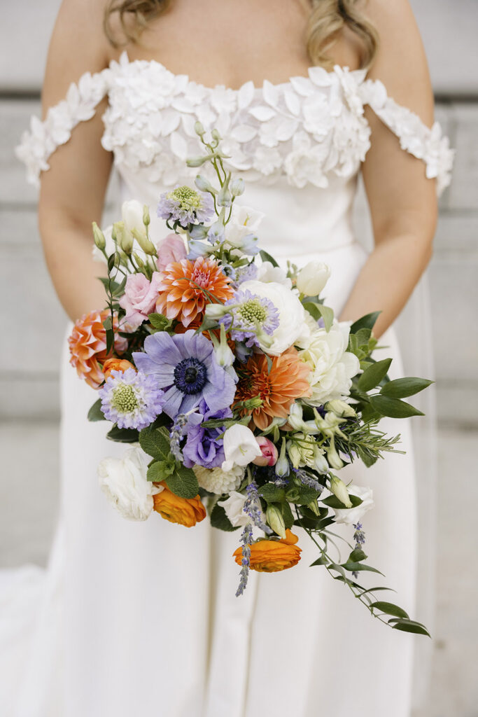 Wedding bouquet with bright orange, lavender, white, and pink flowers held against a textured floral wedding dress.