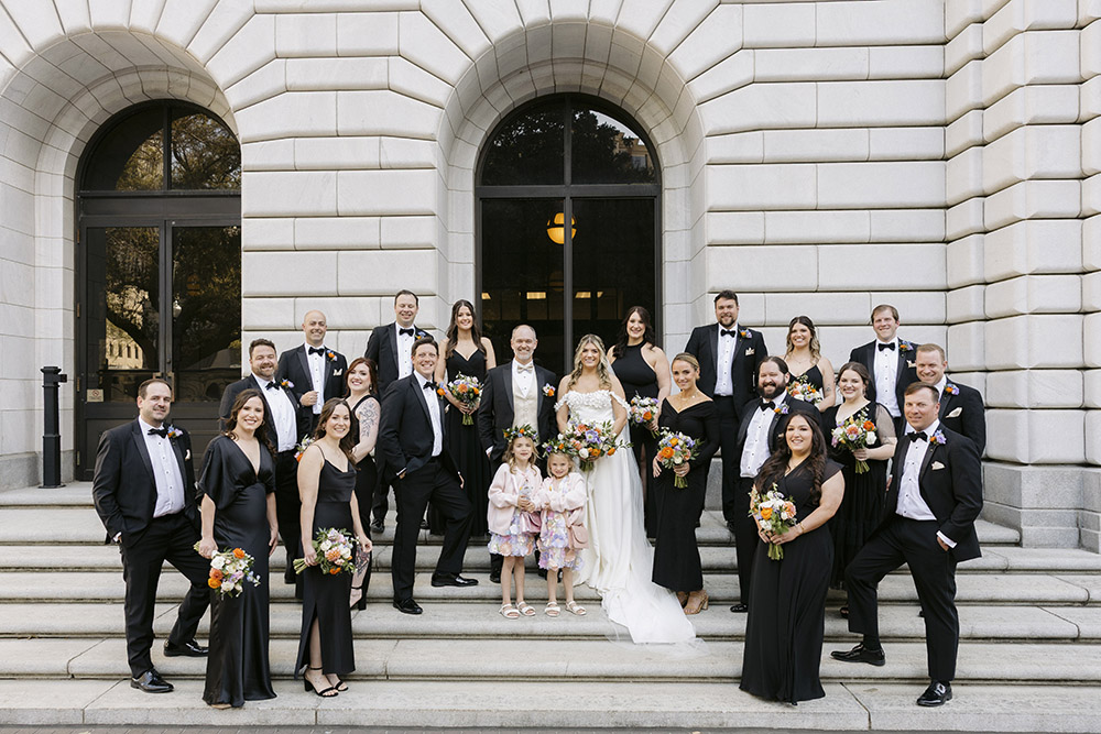 Wedding party portrait on stone steps outside a historic building with attendants in black formal attire holding bouquets.