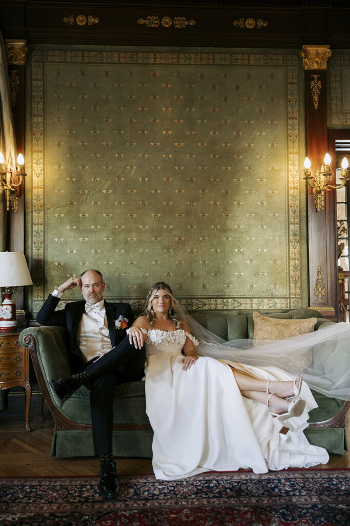 Couple relaxing together on a vintage green sofa inside an ornate historic room during wedding portraits.