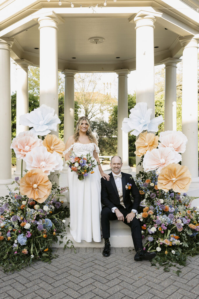 Couple posing in front of a gazebo ceremony backdrop decorated with oversized paper flowers and colorful florals.