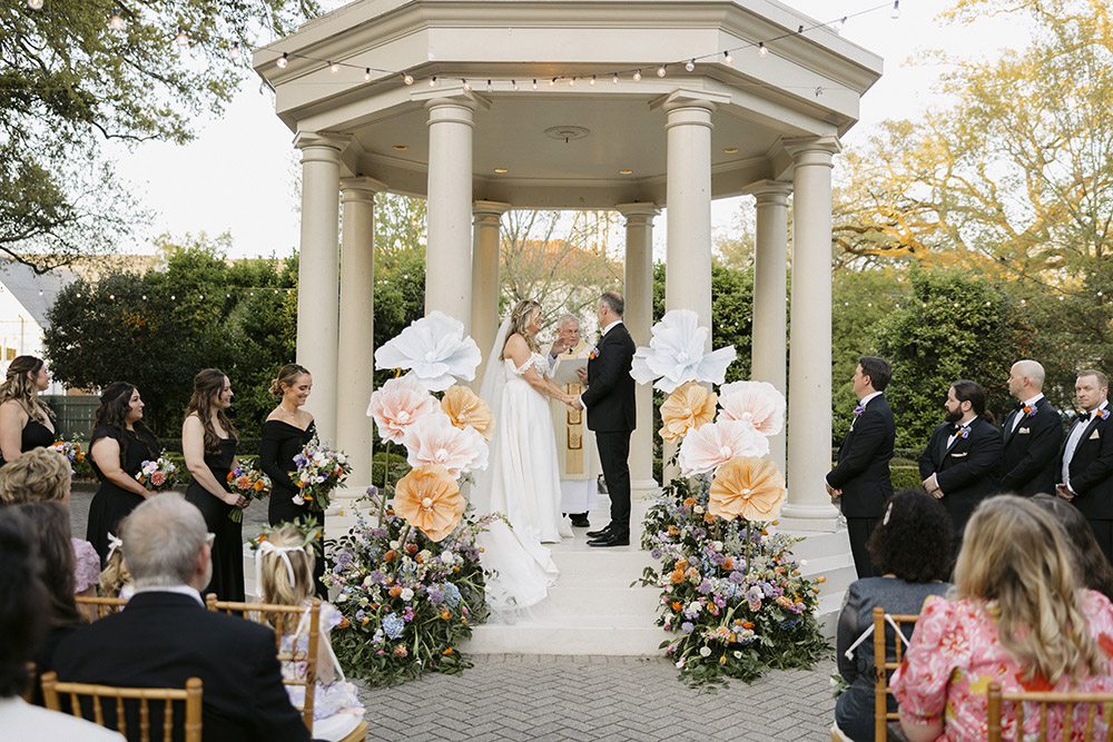 Wedding ceremony moment as couple exchanges vows beneath a columned gazebo with large floral installations.