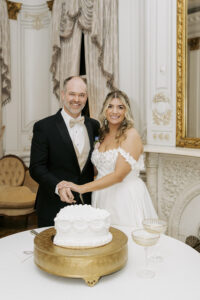 Couple cutting a white wedding cake inside an elegant historic venue with ornate molding and gold mirror.