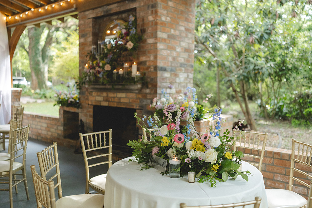 Round reception table with colorful garden-style floral centerpiece beside a decorated brick fireplace.