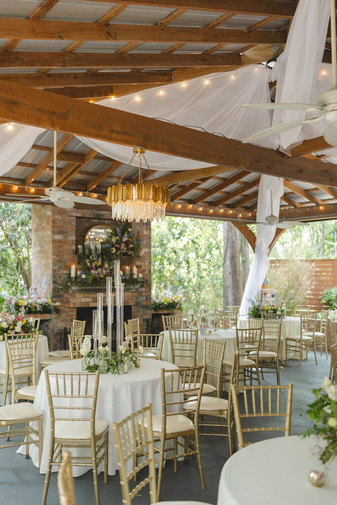Elegant wedding reception setup with gold chiavari chairs, candle centerpieces, and chandeliers beneath draped fabric.