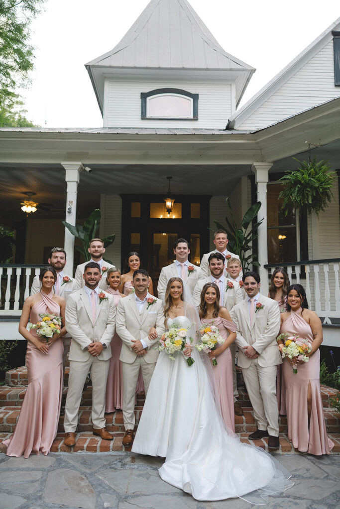 Wedding party portrait on historic home steps with bridesmaids in blush dresses and groomsmen in beige suits.