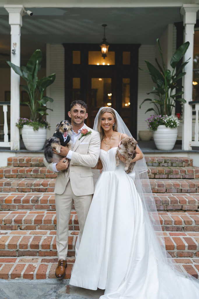 Couple posing with their two dachshunds in tuxedo and dress outfits on the steps of their wedding venue.