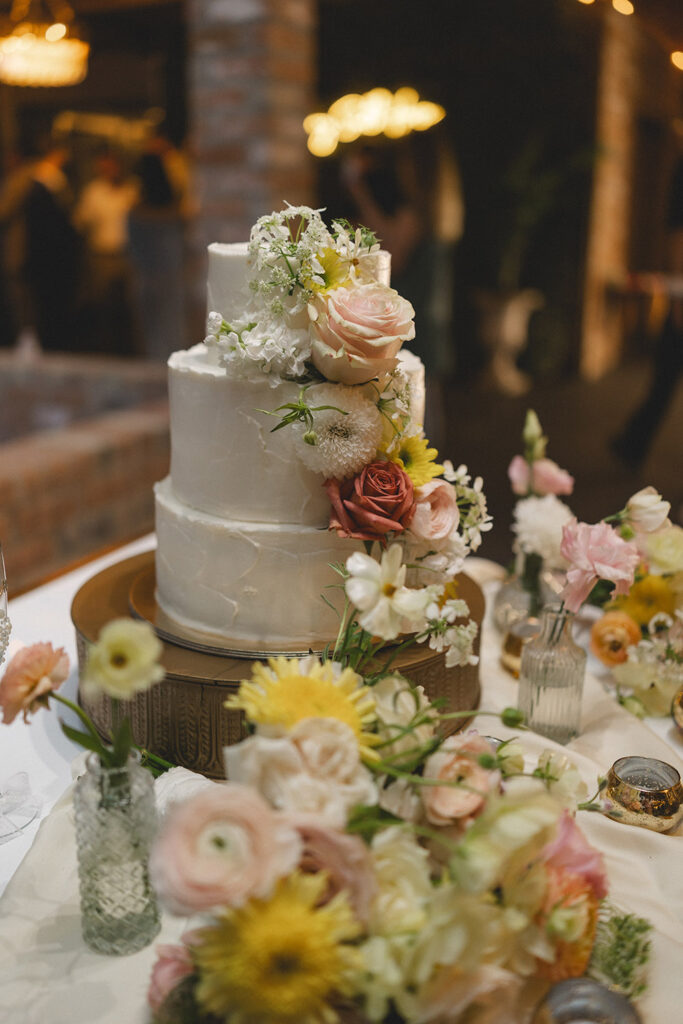 Three-tier white wedding cake decorated with fresh pastel flowers and greenery on a reception table.