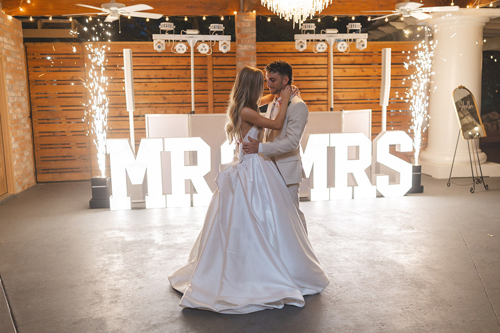 Couple sharing their first dance in front of illuminated “MR & MRS” letters with indoor sparkler fountains.